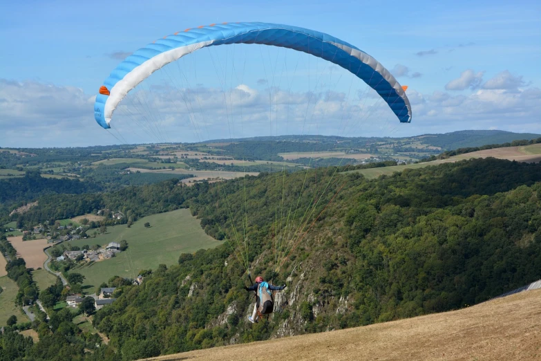 a man flying a kite over a lush green hillside, a picture, by Alison Watt, pulling the move'derp banshee ', aero dynamic, built on a steep hill, long view