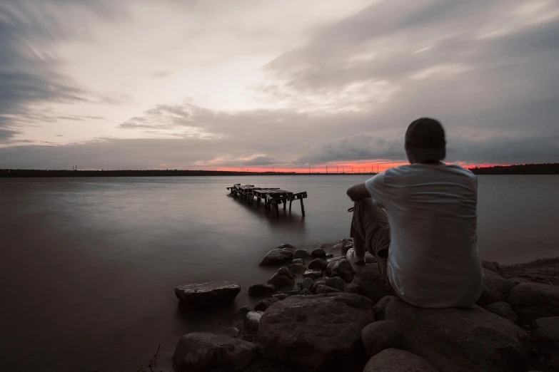 a man sitting on a rock next to a body of water, a picture, by Sebastian Spreng, low sunset, near a jetty, aleksander rostov, self deprecating