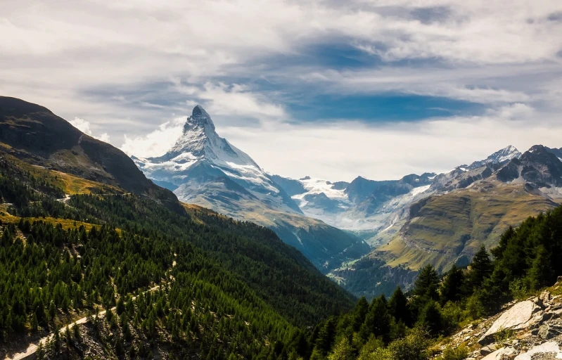 a mountain with a snow covered peak in the distance, a tilt shift photo, by Franz Hegi, shutterstock, art nouveau, in an epic valley, in the swiss alps, cone, gigantic landscape!