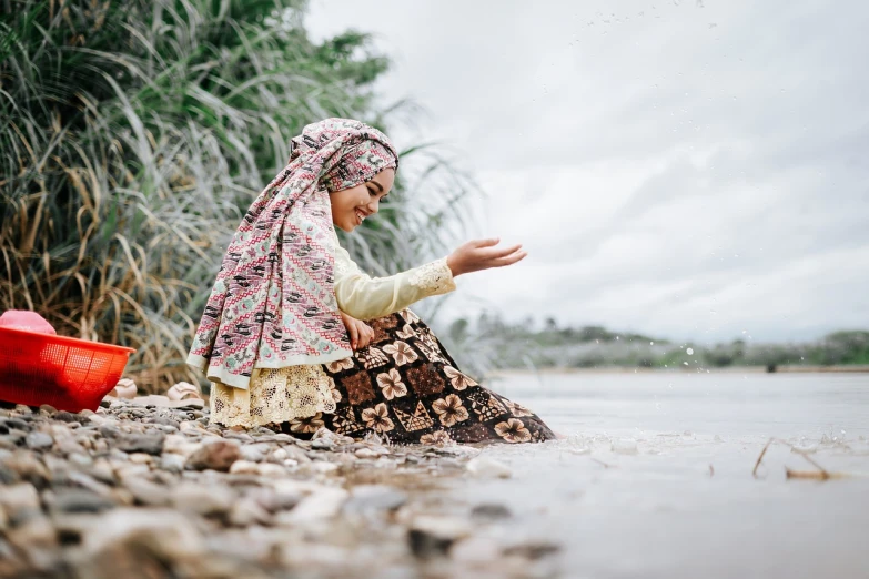 a woman sitting on a rock next to a body of water, by Basuki Abdullah, hurufiyya, petite girl, doing a prayer, wearing a dress made of beads, high resolution photo