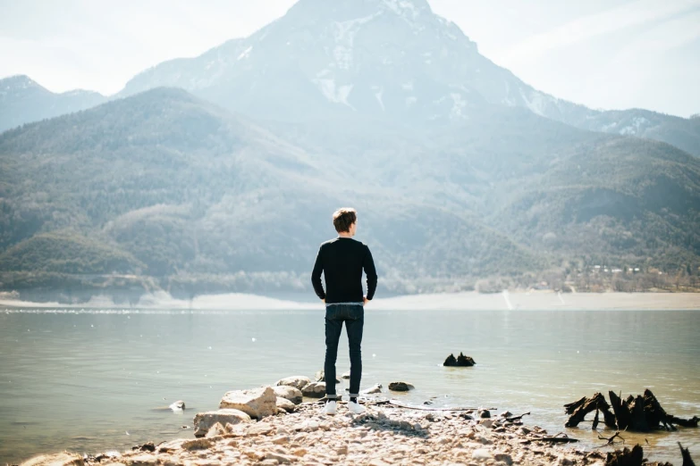 a man standing on top of a rocky beach next to a body of water, a picture, shutterstock, standing in front of a mountain, in full growth from the back, young handsome man, 5 5 mm photo