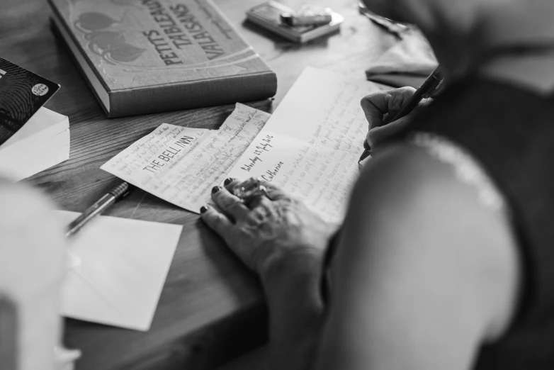 a woman sitting at a table writing on a piece of paper, a black and white photo, by Matthias Stom, pexels, with some hand written letters, celebration, tyler west, morning detail