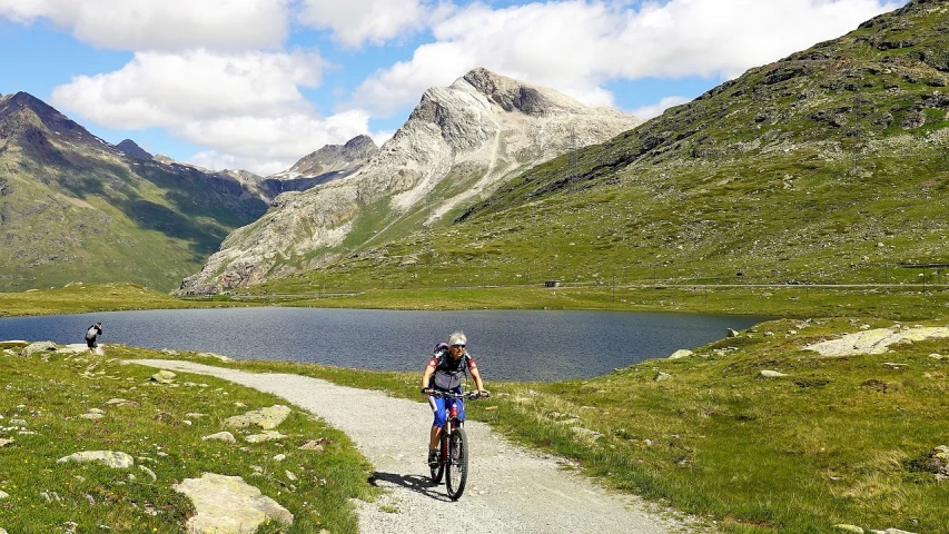 a man riding a bike down a dirt road next to a lake, by Werner Andermatt, pixabay, les nabis, in a mountain valley, female gigachad, simone graci, downhill landscape