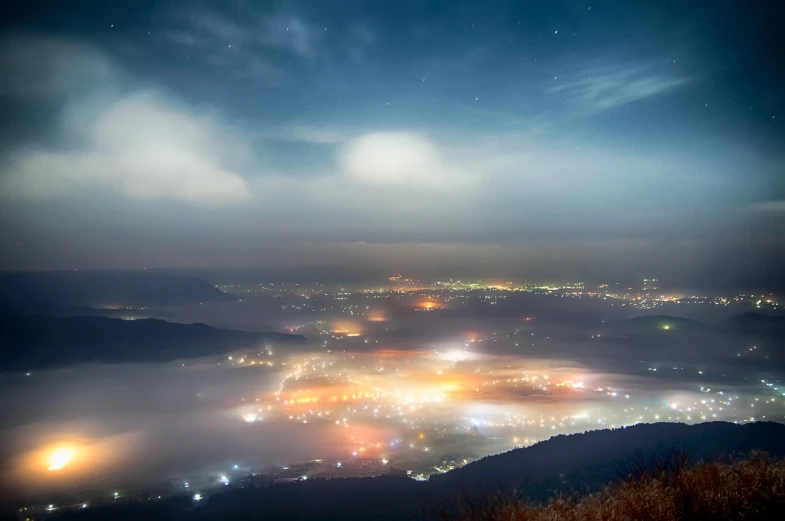 a view of a city at night from the top of a hill, a tilt shift photo, mountains in fog background, long exposure photo