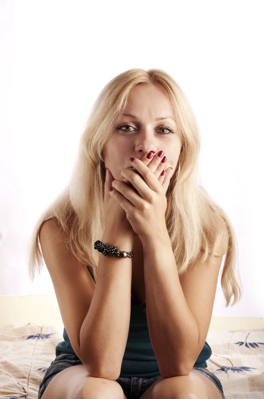 a beautiful blond woman sitting on top of a bed, a stock photo, by Aleksander Gierymski, shutterstock, romanticism, hand over mouth, teeth gritted, sitting with wrists together, compressed jpeg