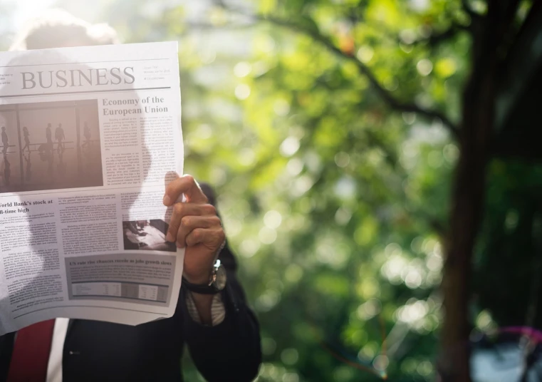 a man holding up a newspaper in front of his face, a picture, unsplash, trees in the background, business surrounding, back - lit, a person standing in front of a