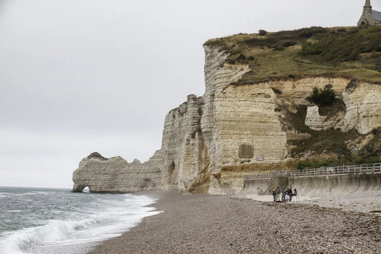 a group of people standing on top of a beach next to the ocean, a picture, by Etienne Delessert, shutterstock, renaissance, chalk cliffs above, overcast weather, france, stock photo