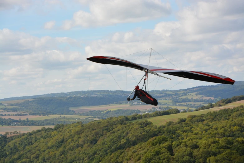 a man flying a hang glider over a lush green hillside, a picture, by Edward Corbett, shutterstock, black mountains, b - roll, flying car, 7000mm film