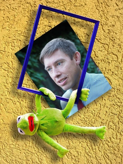 a stuffed frog laying on the ground next to a picture of a man, a photo, inspired by Jan Konůpek, portrait of kermit the frog, digital art!!, ventriloquist dummy, movie frame