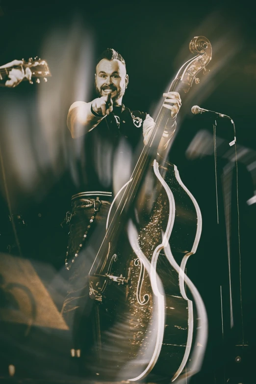 a man standing next to a bass on a stage, a photo, lowbrow, burlesque psychobilly, detailed zoom photo, bearded, instrument of life