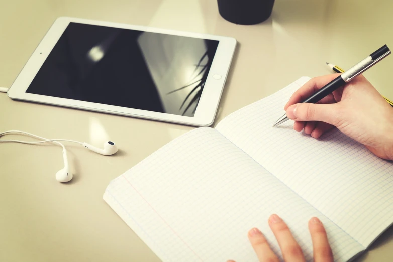 a person writing in a notebook next to a tablet and headphones, a picture, pristine and clean design, discovered photo, compressed jpeg, computer wallpaper