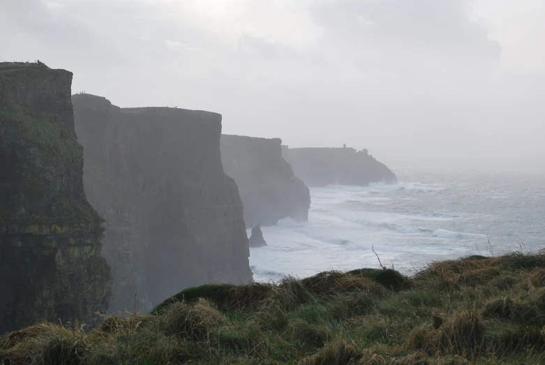 a man standing on top of a cliff next to the ocean, a picture, by Edward Corbett, winter mist around her, extremely strong wind, canyon, clover