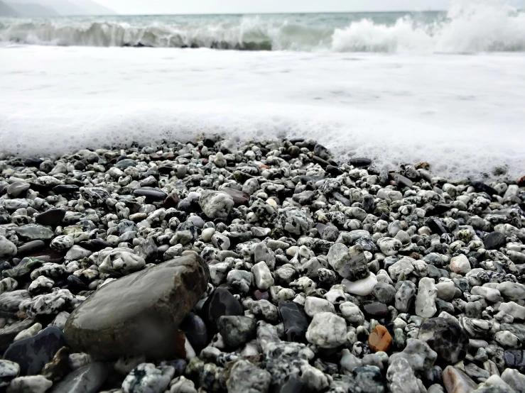 a pile of rocks sitting on top of a beach next to the ocean, a picture, by Milton Menasco, pexels, sea foam, black sea, 8 k. filling of the view, pov photo