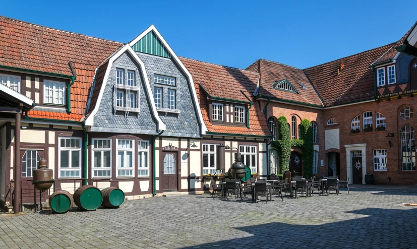 a courtyard with tables and chairs in front of a building, a photo, by Karl Hagedorn, shutterstock, lower saxony, gambrel roof building, wide screenshot, house's and shops and buildings