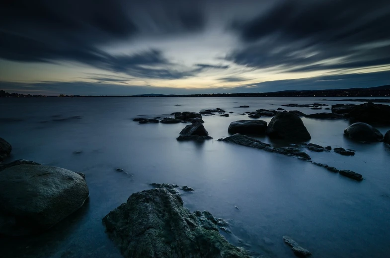 a body of water surrounded by rocks under a cloudy sky, a picture, by Andrew Domachowski, nightfall. quiet, rhode island, dark blue atmosphere, timelapse