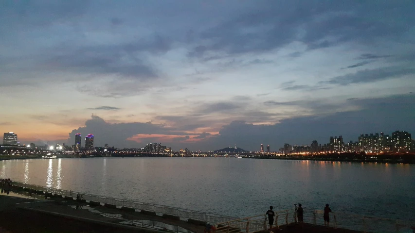 a group of people standing on top of a pier next to a body of water, city twilight landscape, sangyeob park, iphone picture, super wide