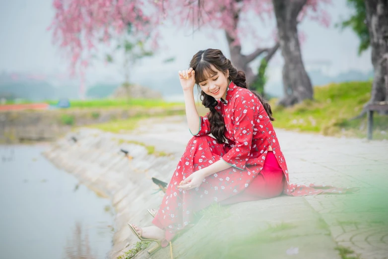 a woman in a red dress sitting next to a body of water, a picture, trending on cg society, shin hanga, falling cherry blossom pedals, with a happy expression, wearing pajamas, 2019 trending photo