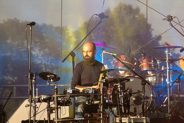 a man that is sitting in front of a drum set, by Dicky Doyle, flickr, against the backdrop of trees, a bald, bartlomiej gawel, tool band