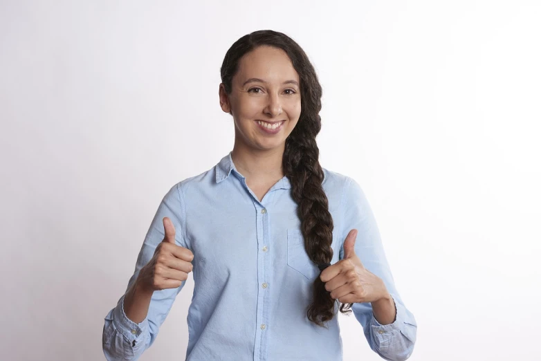 a woman in a blue shirt giving a thumbs up, a stock photo, by Aleksander Gierymski, woman with braided brown hair, on a pale background, high details photo, smiling young woman