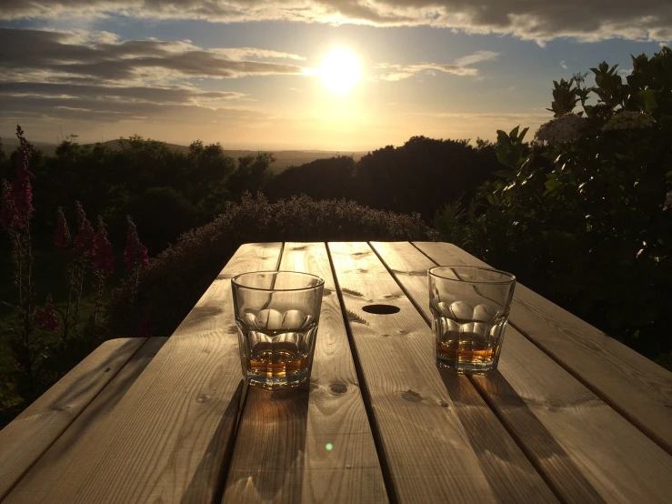 two glasses sitting on top of a wooden table, by John Henderson, romanticism, stunning vista, drinking whiskey, sun glints, cornwall