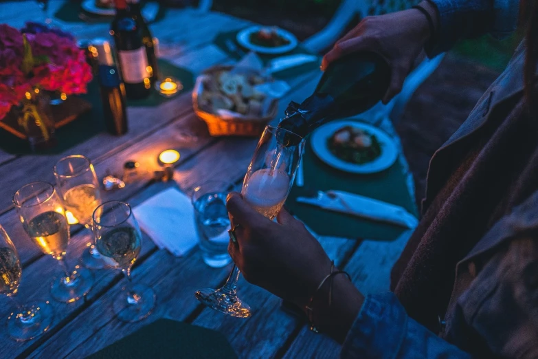 a woman pours a glass of wine at a dinner table, by Niko Henrichon, pexels, night outdoors, holding a bottle of beer, ✨🕌🌙, champagne