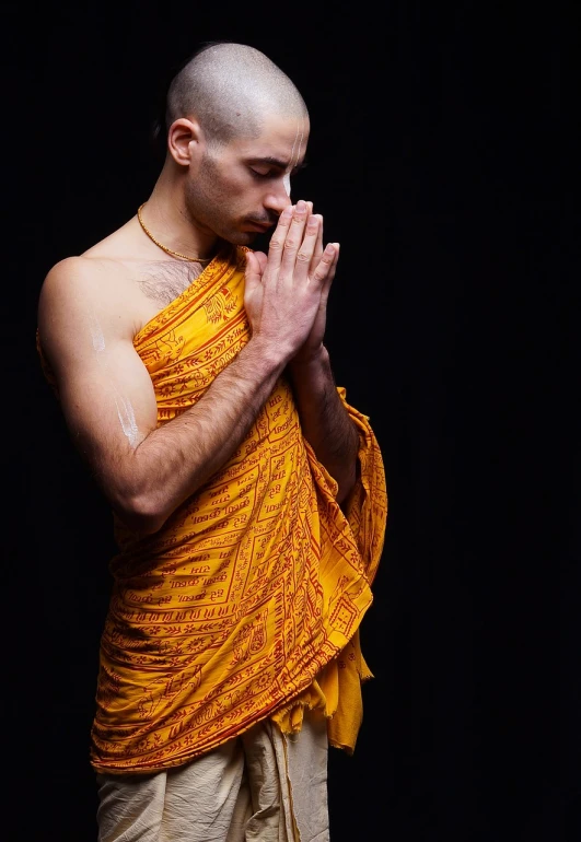 a man standing in front of a black background, a portrait, shutterstock, figuration libre, buddhist monk meditating, dressed in a sari, his palms are sweaty, with yellow cloths