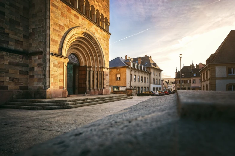 a view of a street with a church in the background, a tilt shift photo, romanesque, setting sun. golden hour, town center background, sweeping arches, low angle dimetric composition