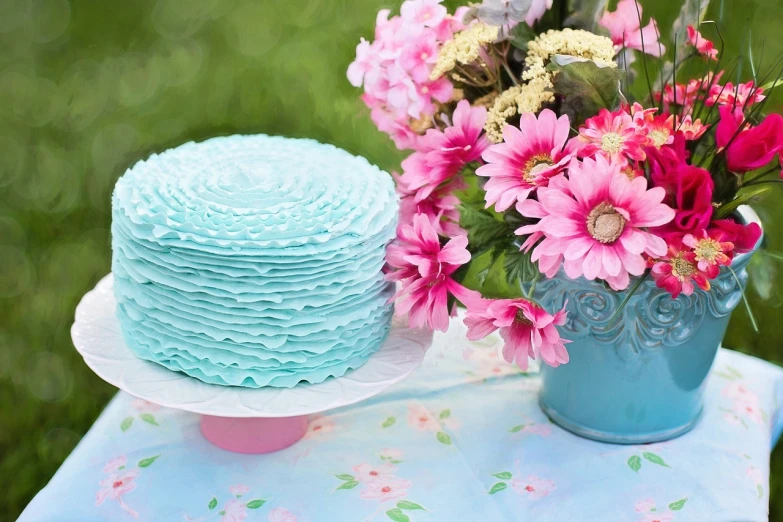 a blue cake sitting on top of a table next to a vase of flowers, a picture, by Rhea Carmi, pexels, turquoise pink and green, ruffled fabric, blue flower field, background image