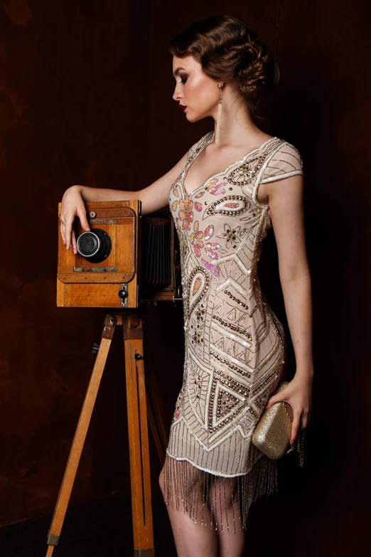 a woman standing next to a wooden camera, a portrait, inspired by Olga Rozanova, cg society contest winner, wearing a sparkling dress, 2 0 s, intricate embroidery, lookbook