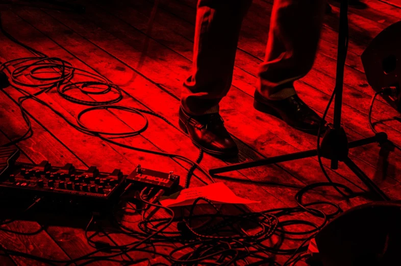 a person standing next to a keyboard on a wooden floor, a picture, by Kurt Trampedach, concert photography, reds, cables, detailed image