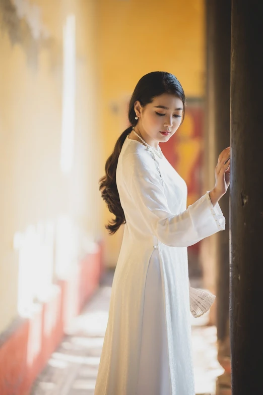 a woman in a white dress leaning against a wall, inspired by Shin Saimdang, shutterstock, ao dai, soft warm light, traditional costume, stock photo