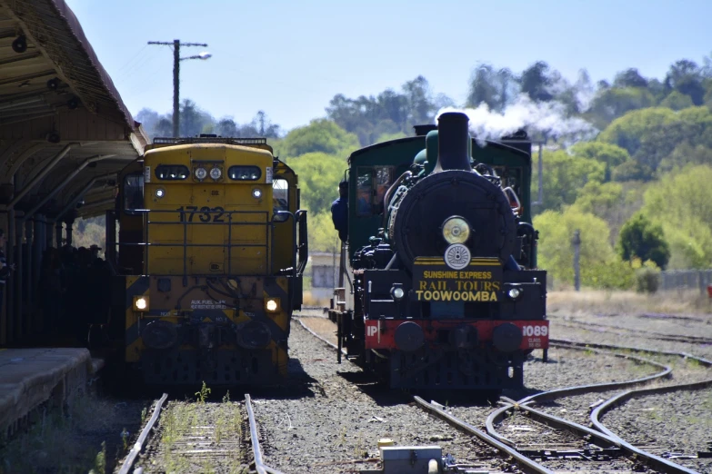 a couple of trains that are sitting on the tracks, by Peter Churcher, tamborine, round about to start, golden engines, sunny day time