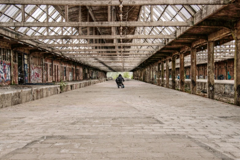 a man riding a skateboard down a cement covered walkway, by Richard Carline, abandoned steelworks, vast empty hall, motorbiker, wooden structures