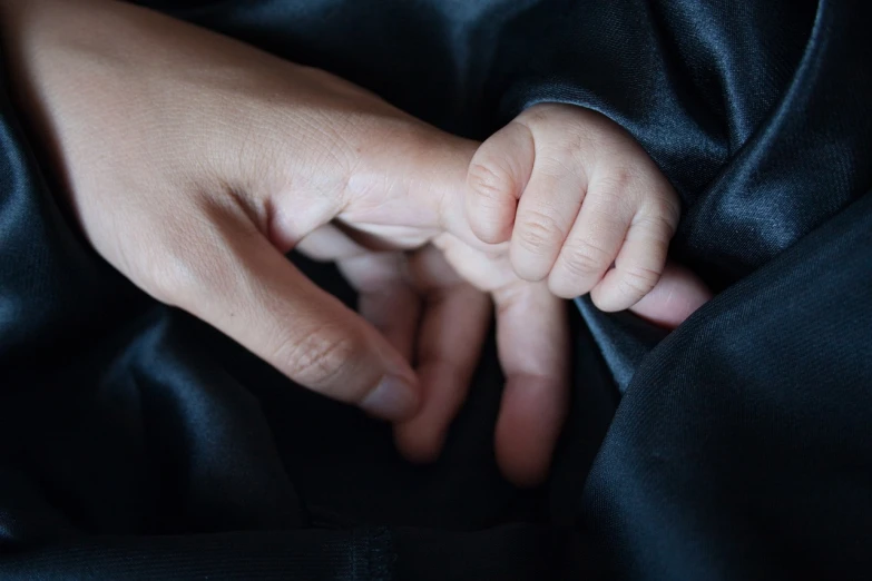 a close up of a person holding a baby's hand, black clothes, curled up under the covers, hand on hip, with index finger