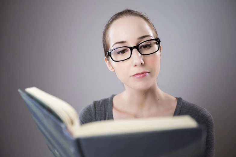 a woman wearing glasses is reading a book, a portrait, academic art, on a gray background, serious focussed look, professional closeup photo