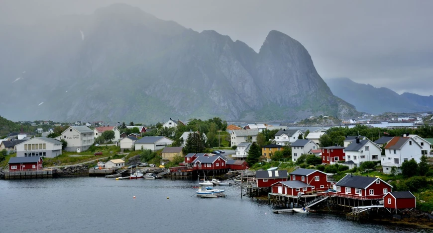 a body of water with a mountain in the background, a picture, by Roar Kjernstad, pixabay, plein air, fishing town, vibrant but dreary red, houses on stilts, july 2 0 1 1