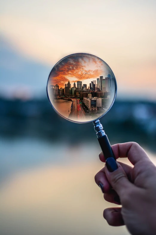 a hand holding a magnifying glass with a city in the background, a picture, realism, reflective lens, superior look, humans exploring, looking at sunset