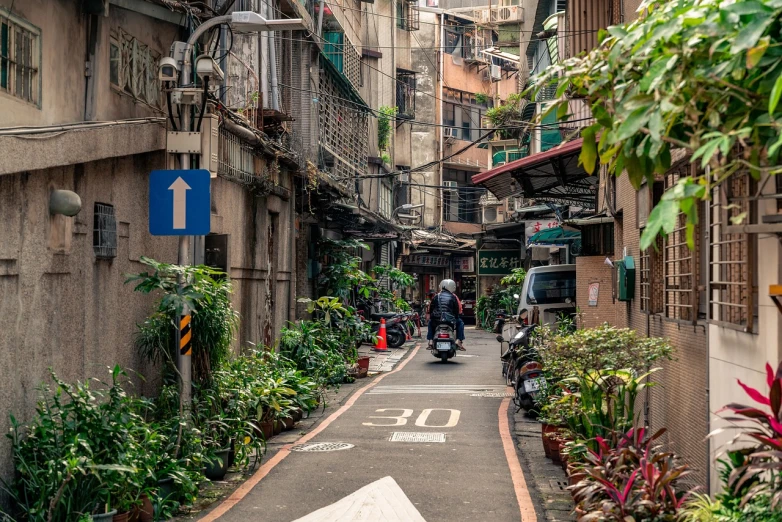 a man riding a motorcycle down a street next to tall buildings, a picture, by Matt Stewart, shutterstock, in a narrow chinese alley, overgrown with lush plants, taiwan, old jeddah city alley