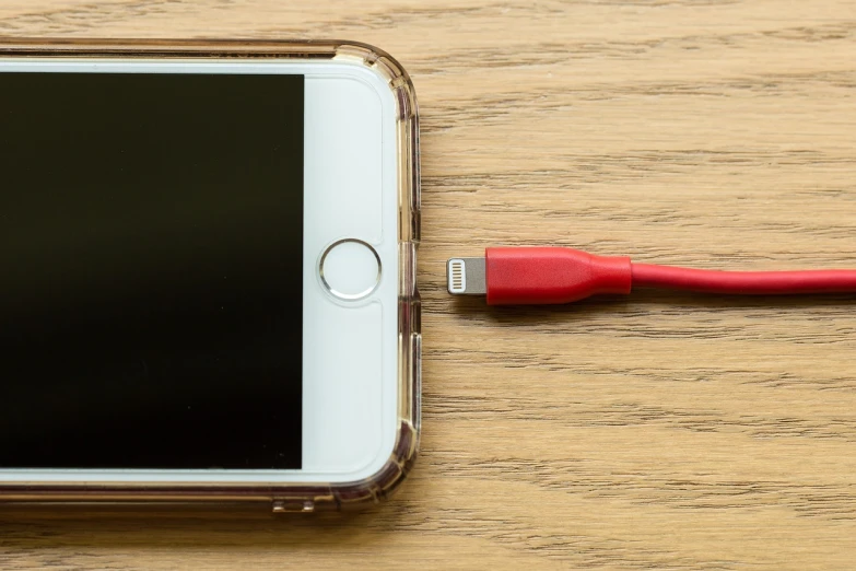 a white cell phone sitting on top of a wooden table, a picture, by Edward Corbett, shutterstock, cable plugged in, red colored, iphone screenshot, battery