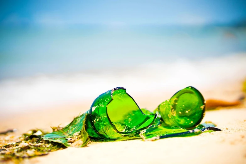 a broken bottle sitting on top of a sandy beach, a stock photo, by Miroslava Sviridova, fine art, green jewelry, glass sculpture, tropical background, closeup - view