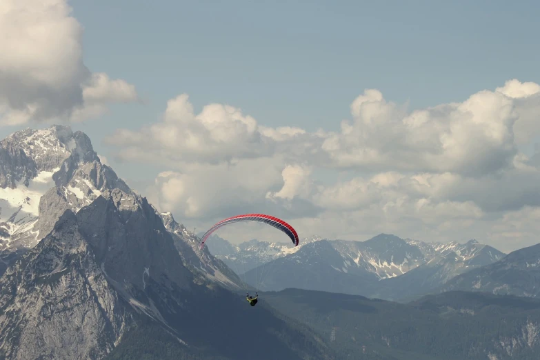 a person paragliding in the mountains on a sunny day, a picture, by Hans Schwarz, shutterstock, wide film still, rocky mountains in background, telephoto shot, deep colours. ”