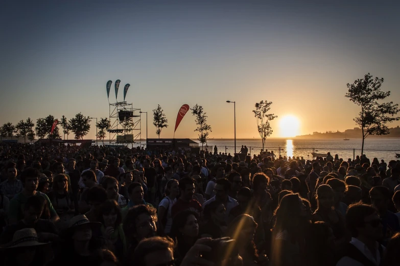 a crowd of people standing on top of a beach, by Joze Ciuha, happening, summer festival in background, sun ray, view from the streets, sunset photo