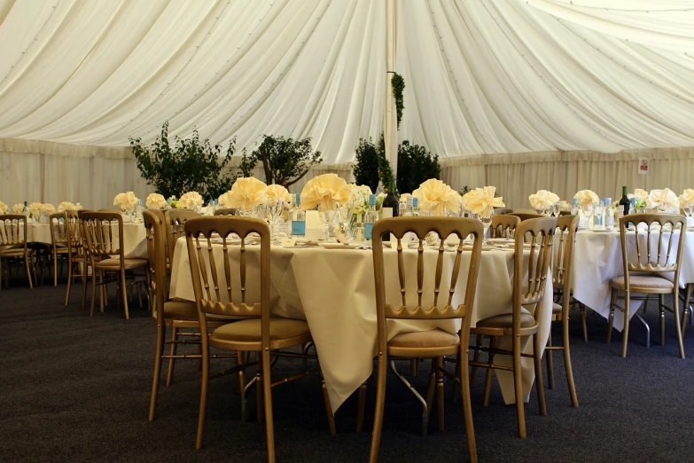 a room filled with lots of tables covered in white cloth, by Joy Garnett, pexels, brown and cream color scheme, tent, accented in bright metallic gold, chairs