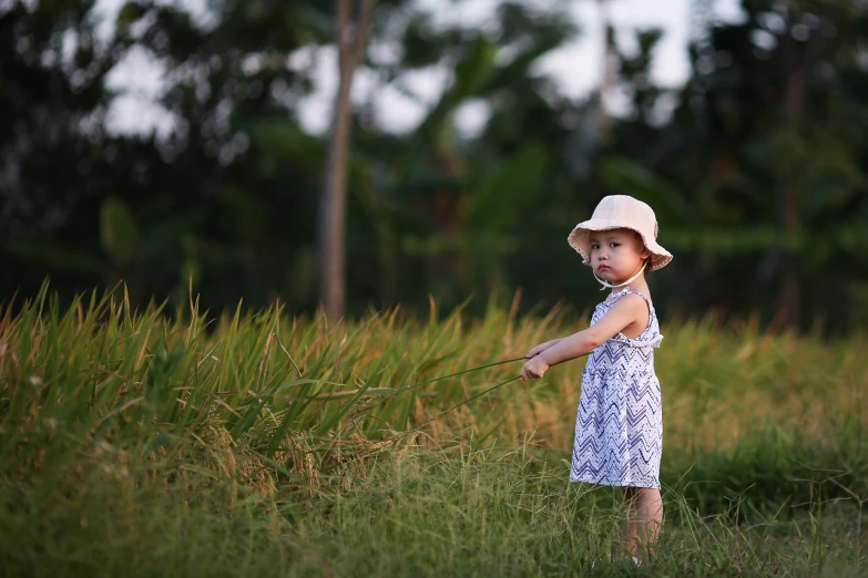 a little girl that is standing in the grass, by Basuki Abdullah, wearing a travel hat, summer evening, bali, 2 years old