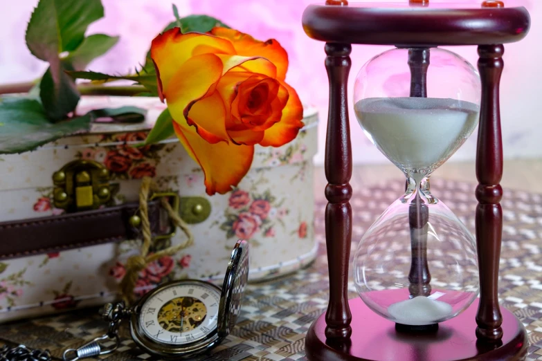 an hourglass sitting on top of a table next to a rose, romanticism, pink and orange colors, clocks, rose background, watch photo