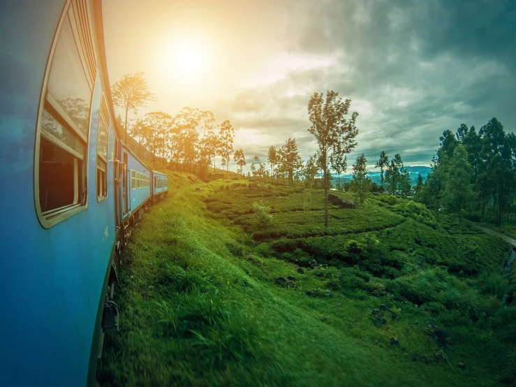 a train traveling through a lush green hillside, by Rodney Joseph Burn, shutterstock, sri lankan landscape, sun set, tones of blue and green, train station background