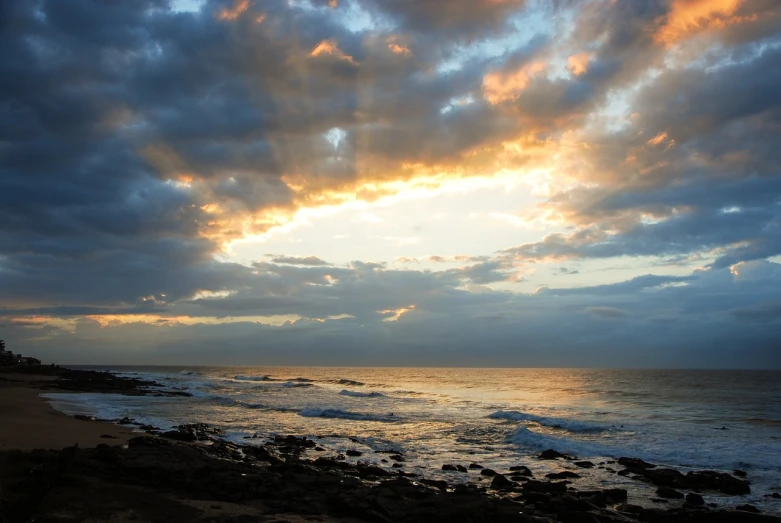 a man riding a surfboard on top of a sandy beach, a photo, by Alexander Scott, romanticism, sunrays breaking through clouds, south african coast, sunset photo, rocky shore