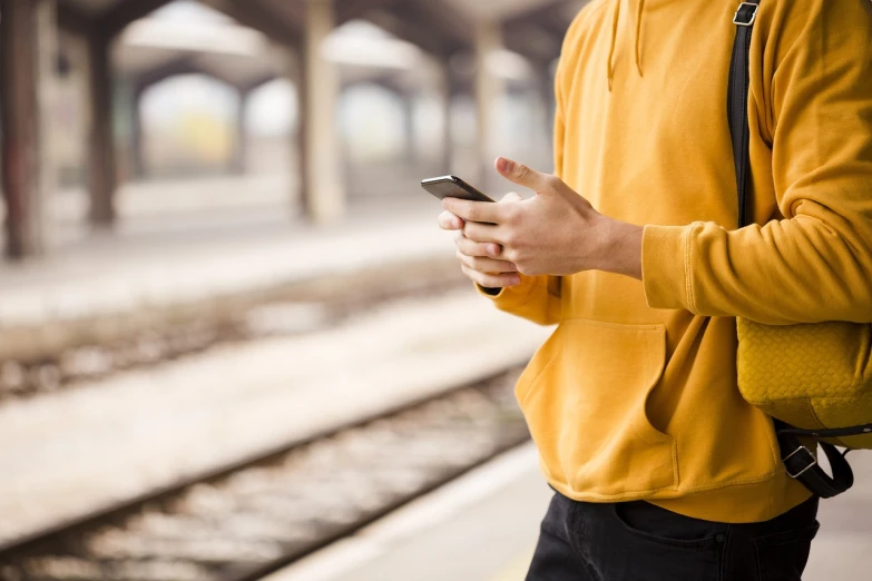 a man standing on a train platform holding a cell phone, shutterstock, wearing a yellow hoodie, high res photo, close-up photo