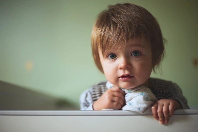 a close up of a child in a crib, by Emma Andijewska, pexels, short brown hair and large eyes, sitting on edge of bed, sad green eyes, fierce expression 4k