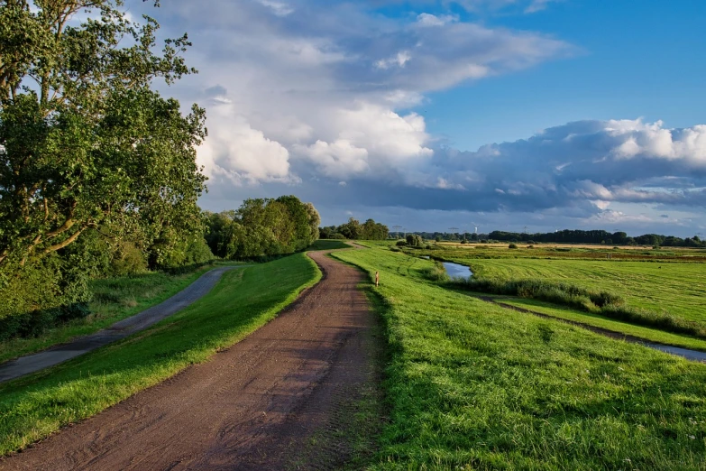 a dirt road running through a lush green field, a picture, by Jacob Esselens, pixabay, realism, canal, late summer evening, low dutch angle, winding rivers